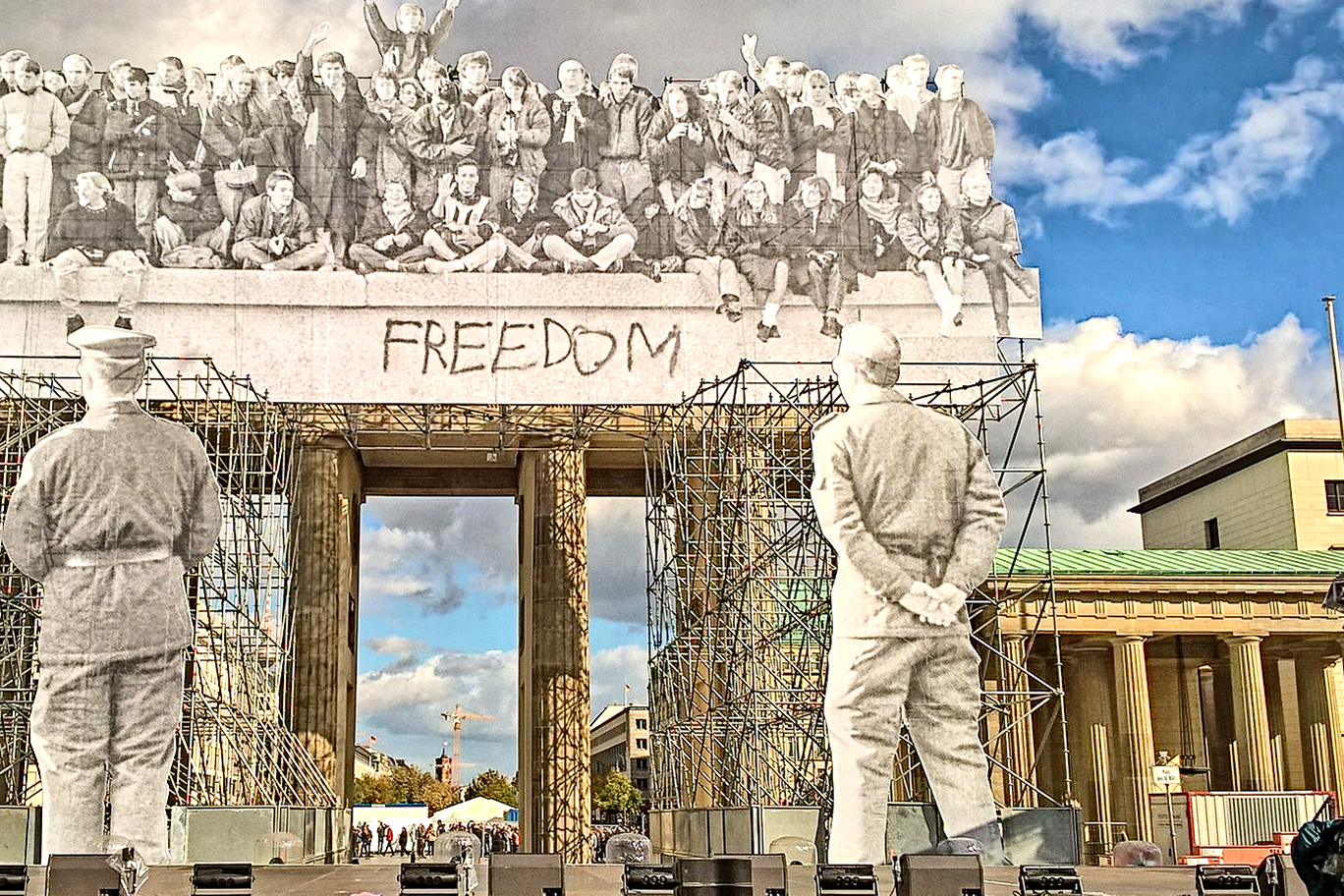 Eine Installation am Brandenburger Tor macht Wandel und Veränderung deutlich. Mittels Gerüsten sind Bilder vor das Tor montiert. Oben auf dem Tor sind Menschen zu sehen, die auf der Berliner Mauer sitzen. Vor dem Tor stehen Grenzsoldaten, die auf die Mauer blicken. Und der Blick des Betrachters führt durch das offene Tor durch.
